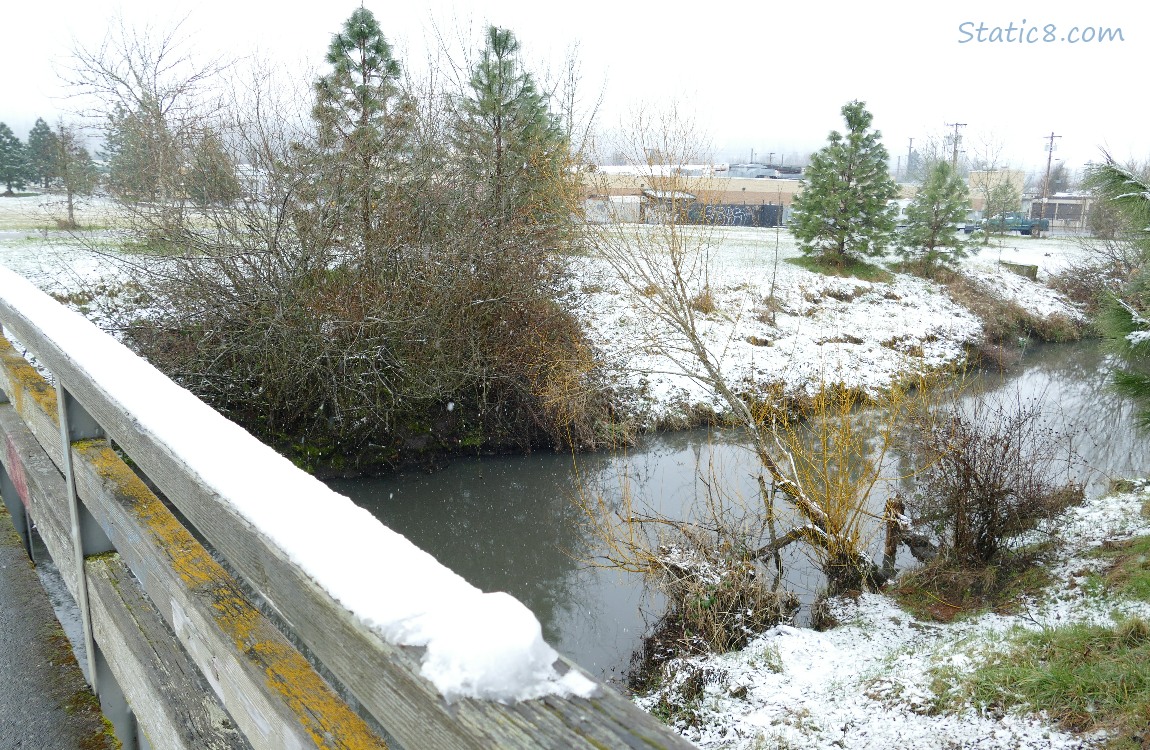 snow along the creek and covering the railing of a walking bridge