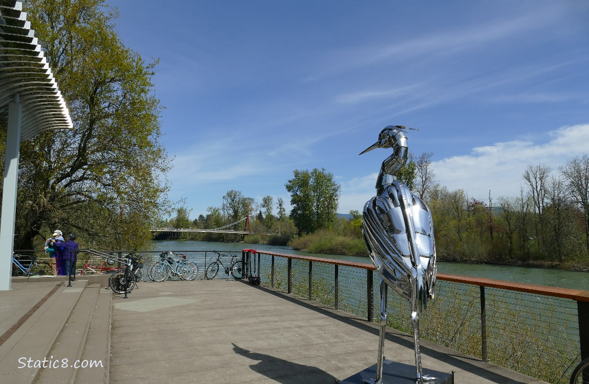 Chrome Heron statue next to a river with a suspension bridge in the distance