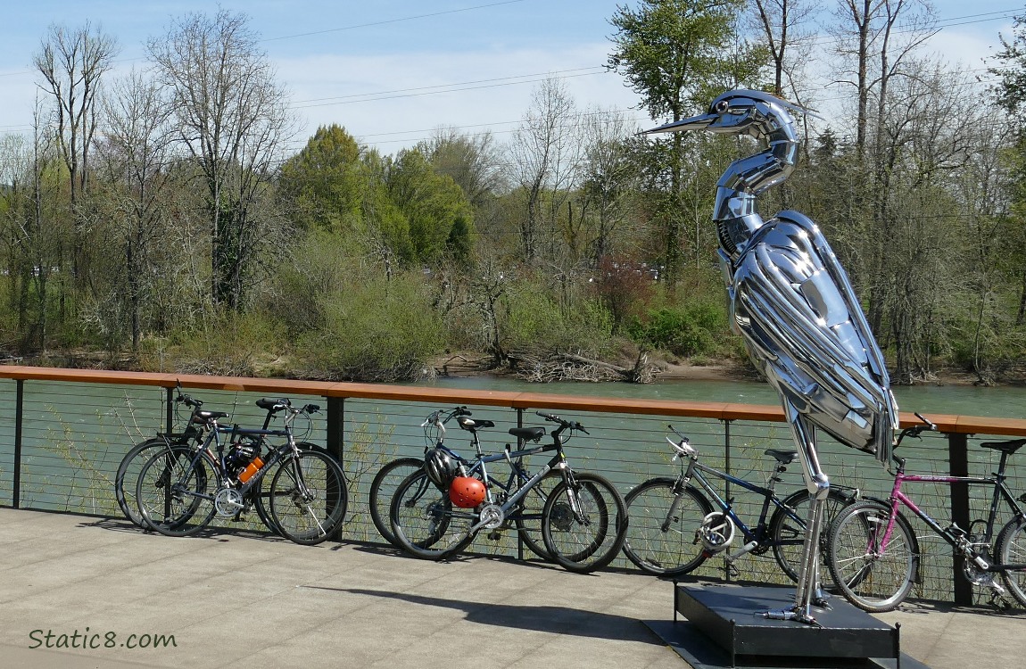 Chrome Heron statue in front of the river, with bikes along the railing and trees on the other side of the river