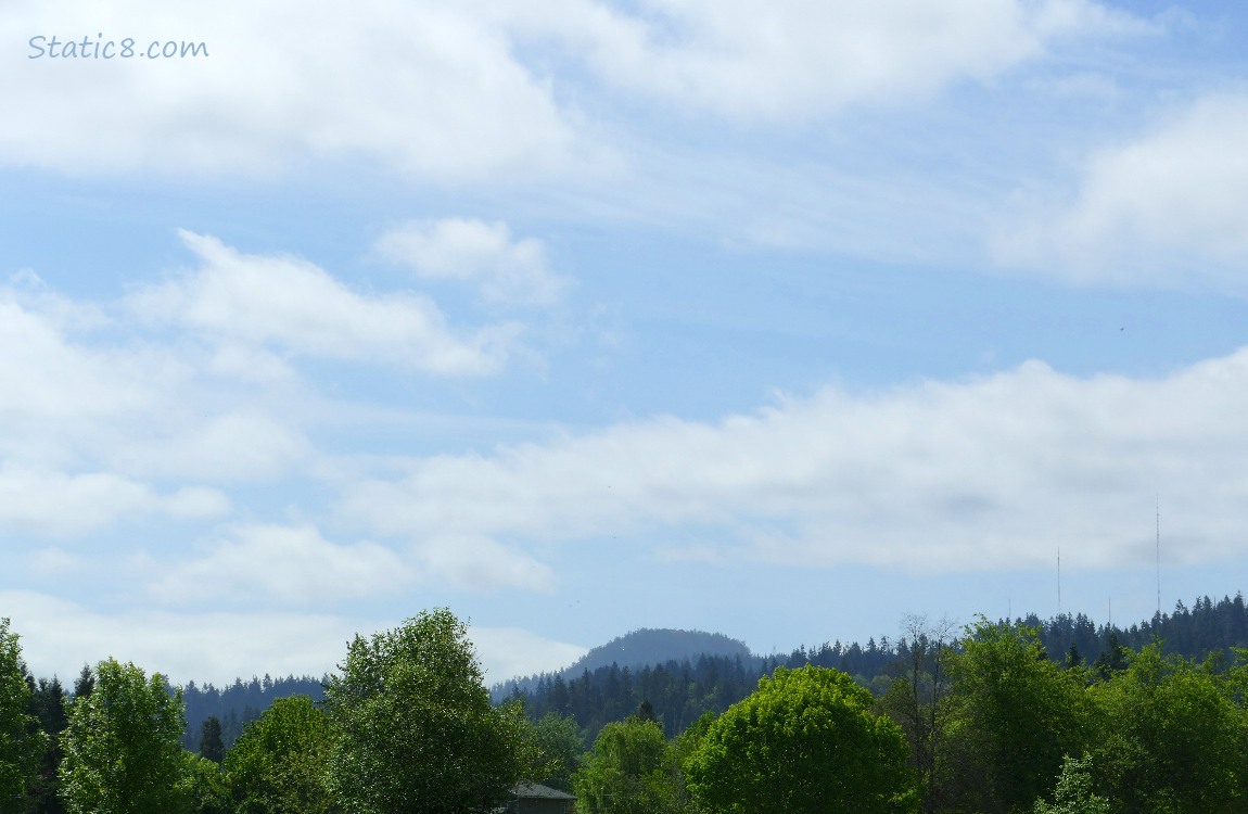 white clouds and blue sky over a hill and trees in the distance